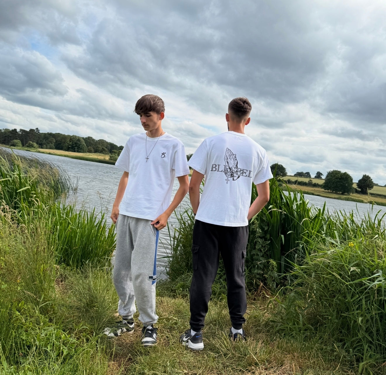 Two models standing by a lake with grass and trees in the background